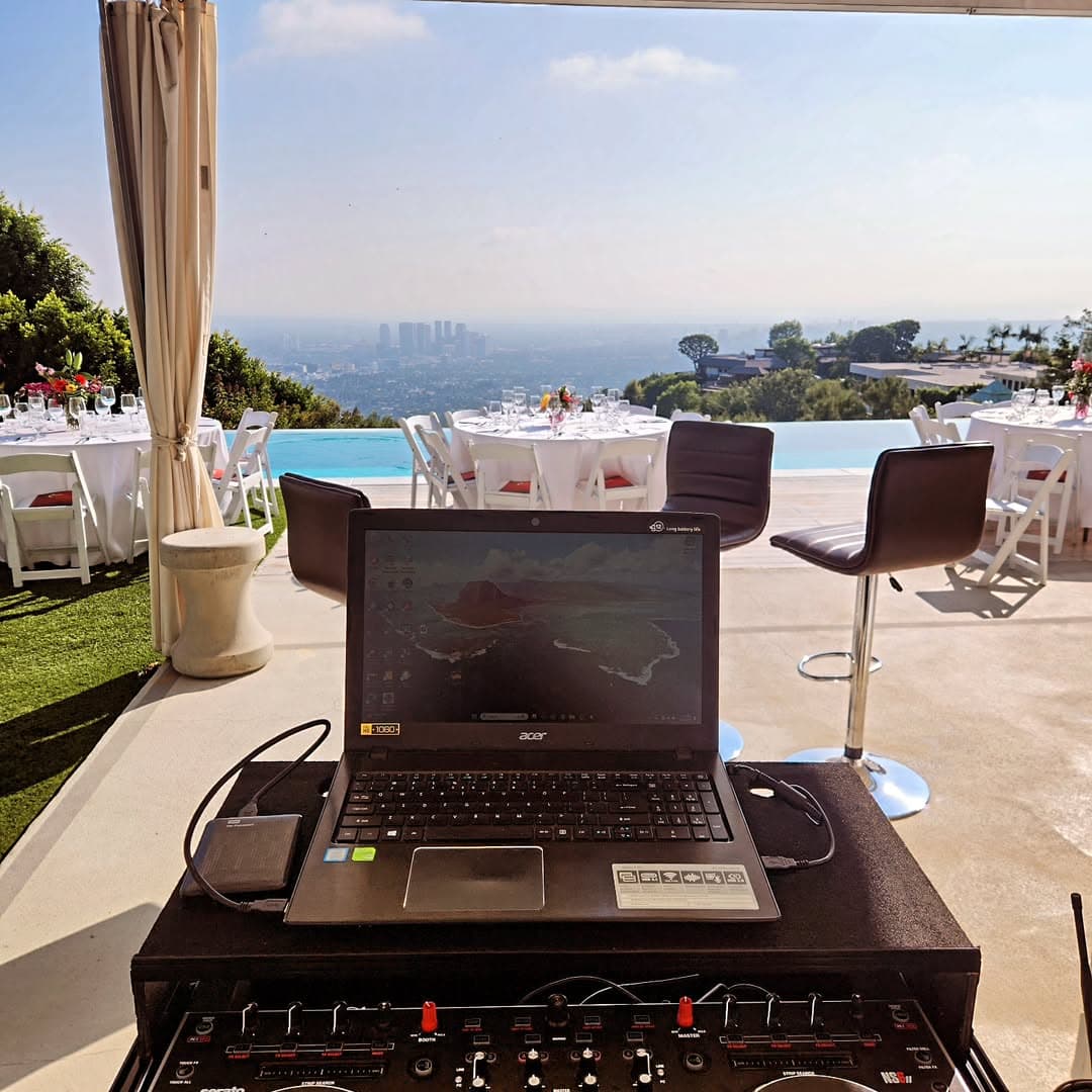 DJ setup overlooking a poolside event with round tables, chairs, and city skyline in the background.