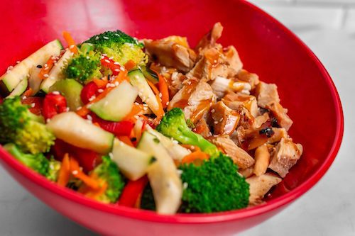 A red bowl filled with sliced cooked chicken and a mix of steamed vegetables, including broccoli, carrots, zucchini, and red bell peppers, topped with sesame seeds.