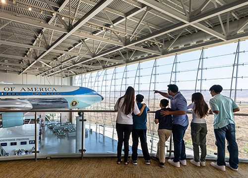 A family observes a large plane model inside a spacious, glass-walled museum. The father points out details as they stand near a railing.