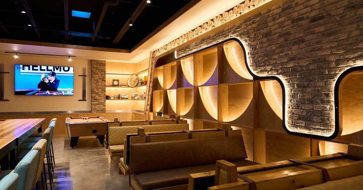 Modern lounge with wooden benches, a pool table, and a TV mounted on a brick wall; illuminated shelves and exposed ceiling create a contemporary atmosphere.