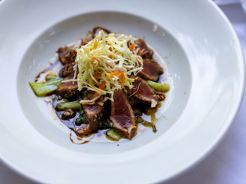 A plate of seared tuna slices on stir-fried vegetables, topped with shredded coleslaw, served with a drizzle of sauce.