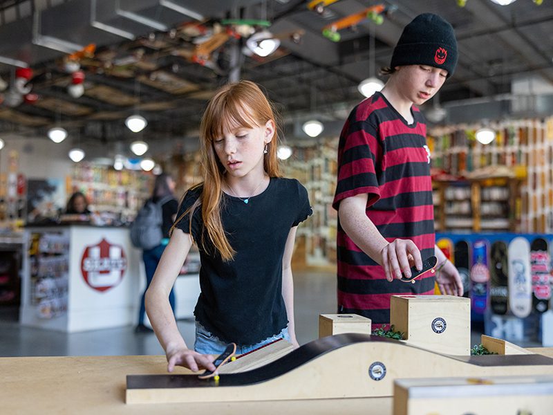 Two children play with fingerboards on wooden ramps inside a skateboard shop, with various skateboards displayed in the background.