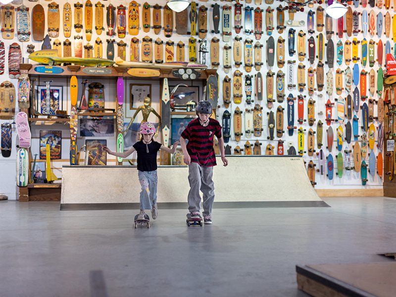 Two children wearing helmets skateboard indoors. The background features a wall covered with a variety of skateboard decks.